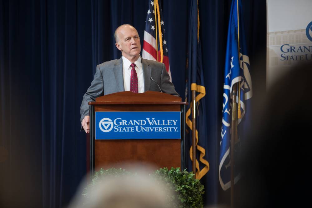 Dean Potteiger talking at a GVSU podium in focus, people's head out of focus in the foreground